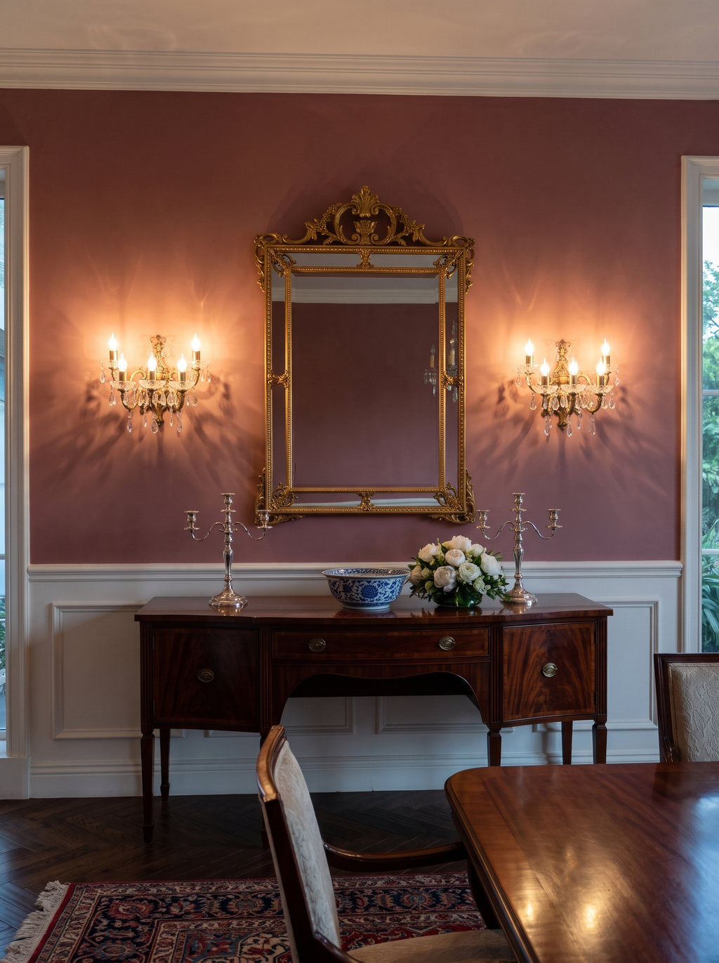 A pair of Bohemia wall lights lit either side of a gilt mirror above a mahogany sideboard in a rose-walled dining room
