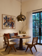 Brass Petals Pendant, Small, lit over a round walnut dining table in a cream-walled farmhouse dining nook with cane chairs and an abstract painting