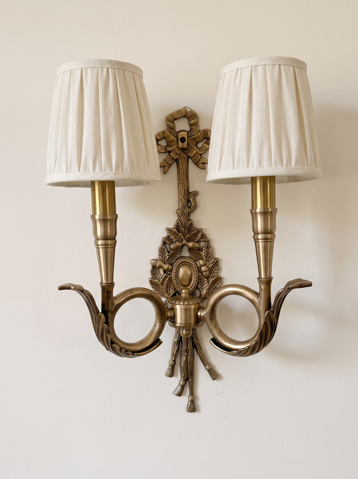 Chasse Cast Brass Double Wall Light, antique brass hunting-horn arms, ribbon bow and oak-leaf trophy with pleated linen shades, front view