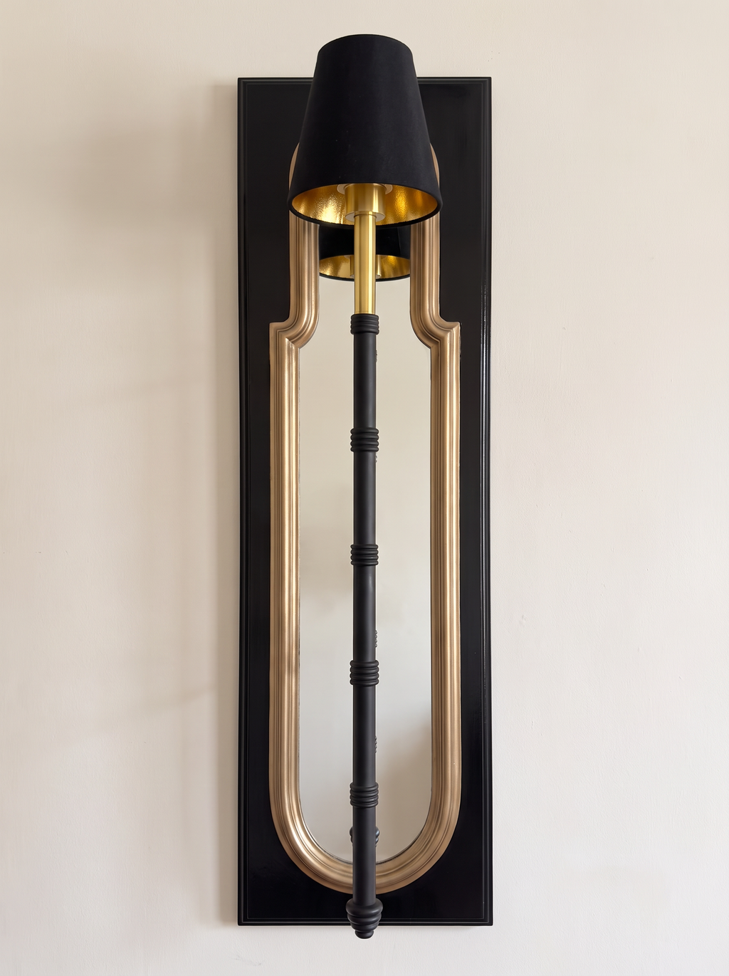 Darpan Mirror Wall Lamp, front view: glossy black panel, gold-framed arched mirror and a black and brass lamp with a black shade
