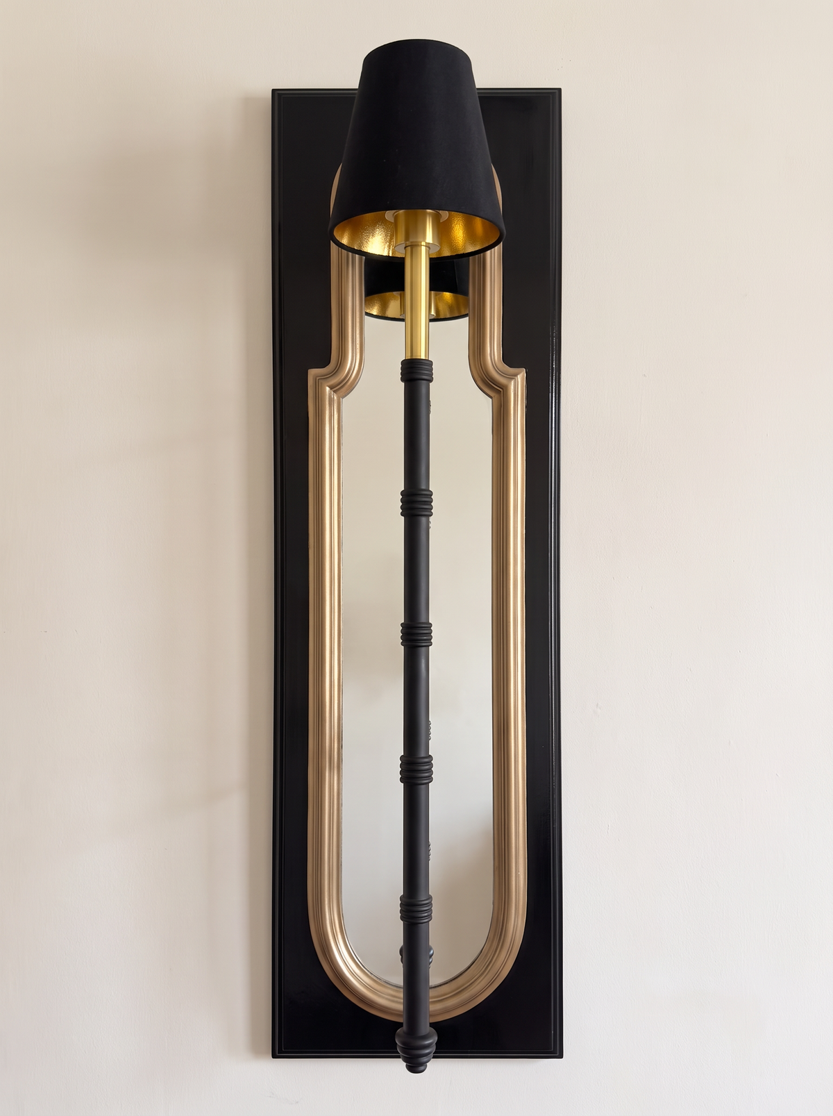 Darpan Mirror Wall Lamp, front view: glossy black panel, gold-framed arched mirror and a black and brass lamp with a black shade