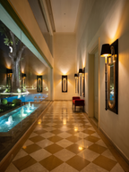 Darpan Mirror Wall Lamps lit along a hallway with checkerboard marble floor and a glass wall onto a lit pool at night