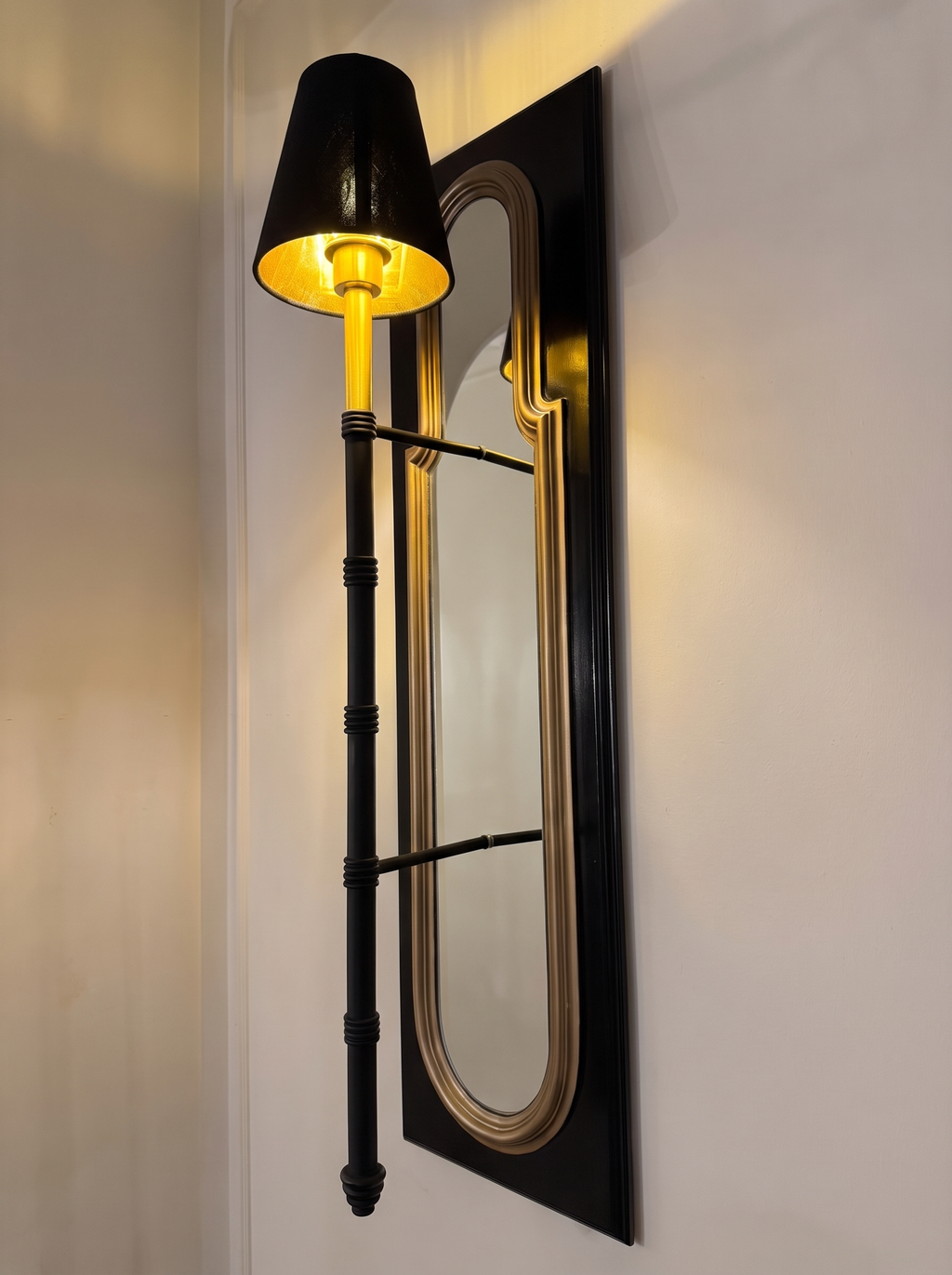 Darpan Mirror Wall Lamp switched on, three-quarter view showing the lamp standing off the mirror on two black arms
