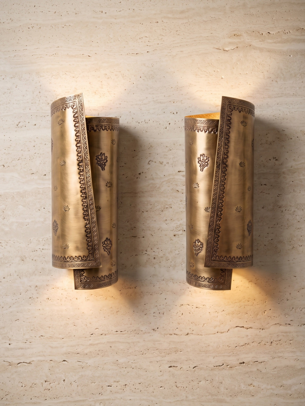 A mirrored pair of Patra Scroll Sconces in hand-hammered brass, glowing up and down on an ivory travertine wall