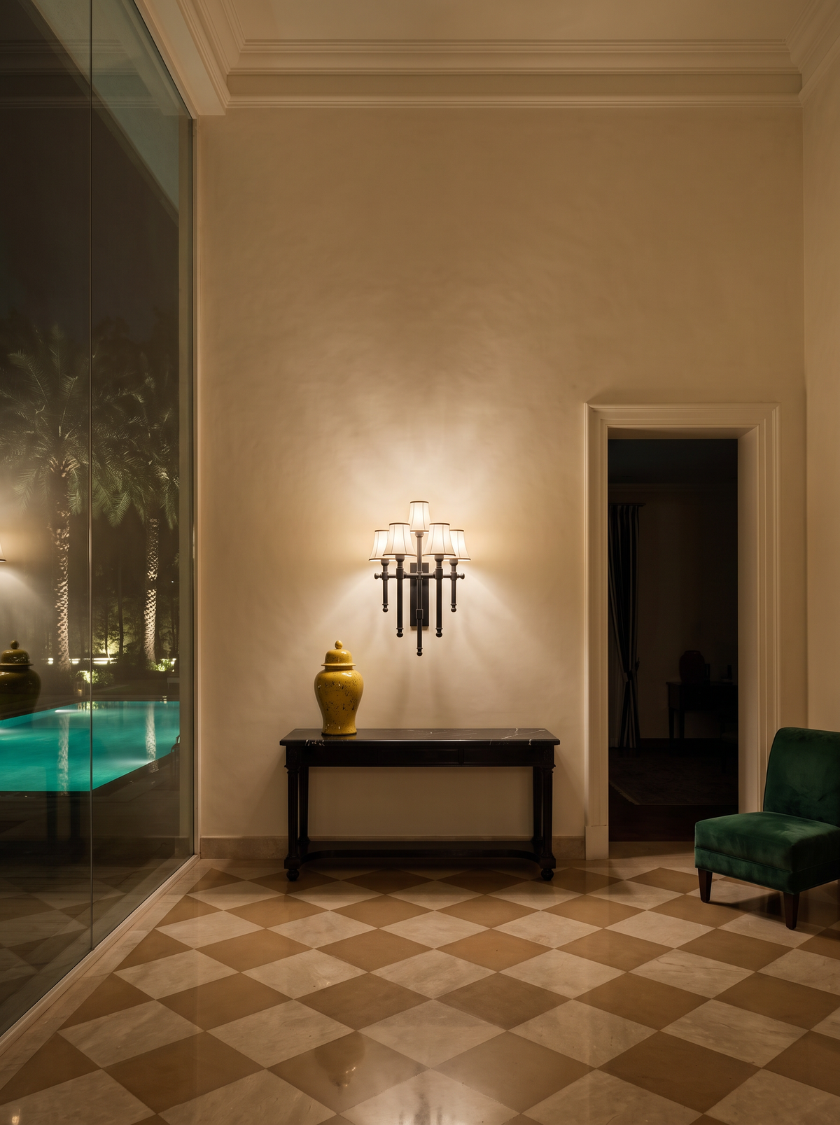 Flambeau Five-Light Wall Light glowing above a black marble console in a tall cream plaster entrance hall at night, with a lit pool beyond the glass and an emerald velvet chair