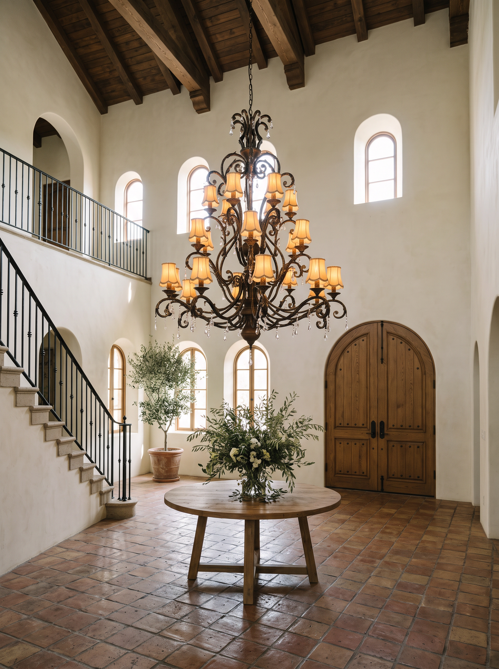 Toledo chandelier lit above a round oak table in a double-height Mediterranean villa hall with timber beams, terracotta floor and arched doors