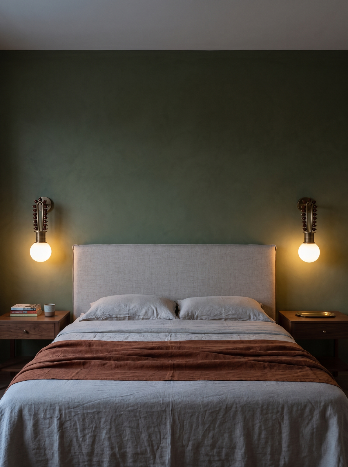 A pair of Mala wall lights glowing on either side of a linen headboard against an olive-green wall