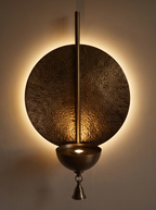 Prabha Halo Wall Light switched on, a warm halo glowing on the wall behind the beaten brass disc