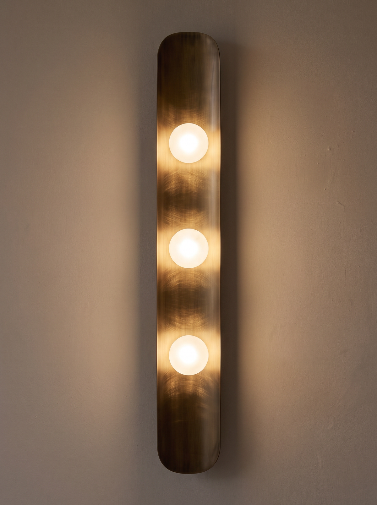 Reed Wall Light switched on, three frosted globes glowing and warm light running down the curved brass