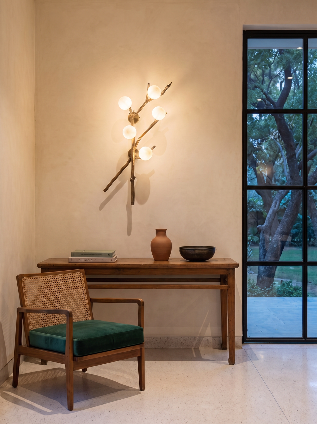 Shakha Branch Wall Light lit above a teak console in a cream-walled farmhouse entrance, with a cane chair and garden view