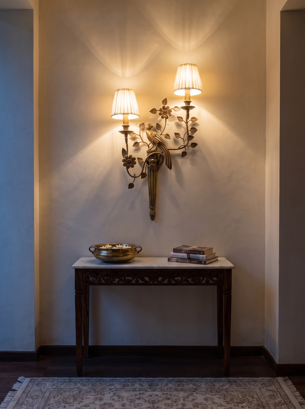 Shuka Double Wall Light lit above a carved console with a brass urli in a foyer at dusk