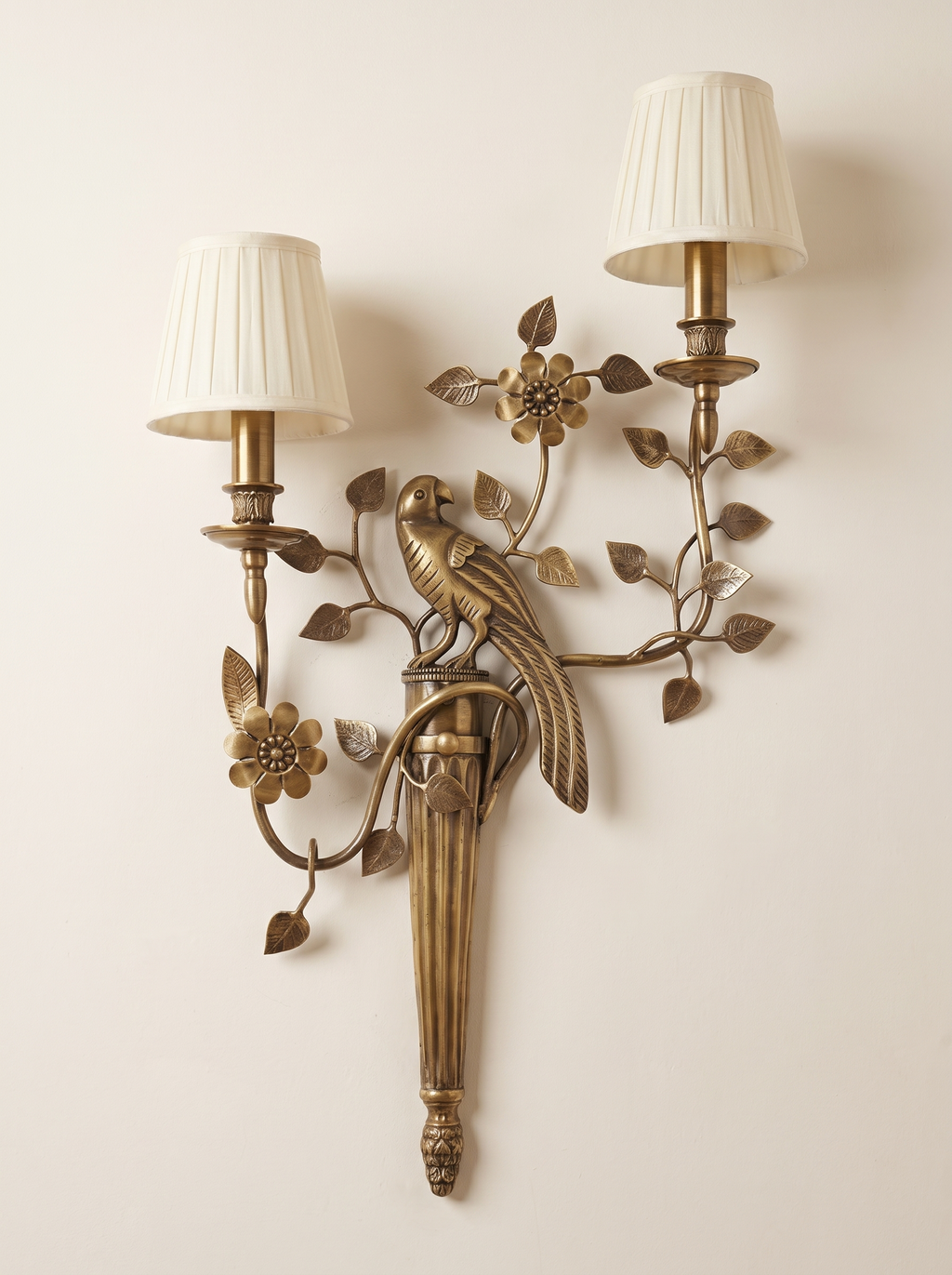 Shuka Double Wall Light in antique cast brass with a parrot, leafy branches and two pleated ivory shades, front view