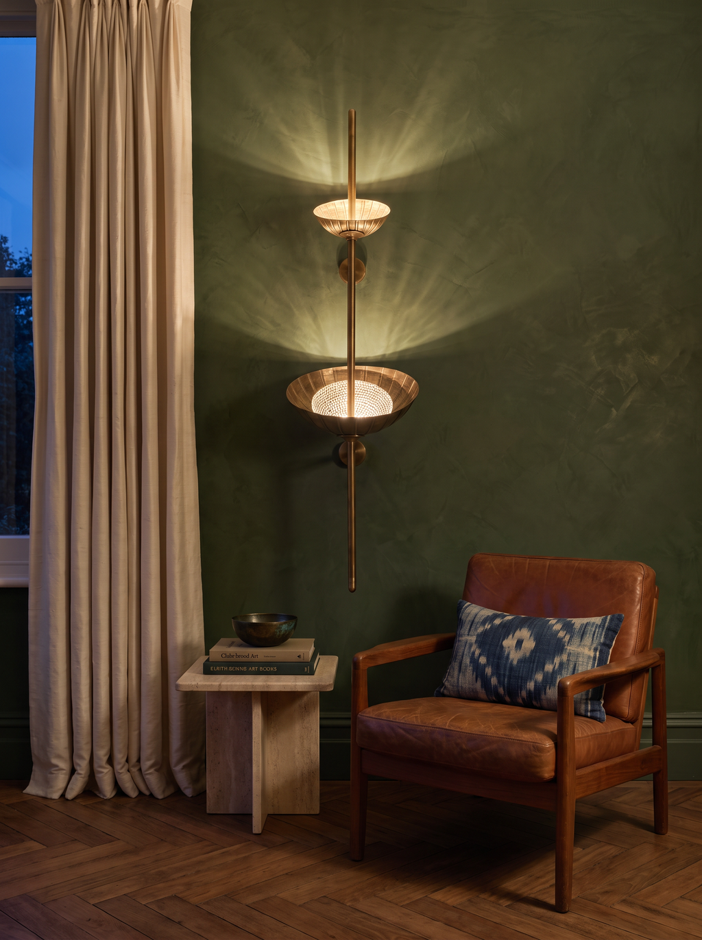 Stambha Etched Brass Wall Light glowing on a moss-green Venetian plaster wall beside a cognac leather easy chair at dusk