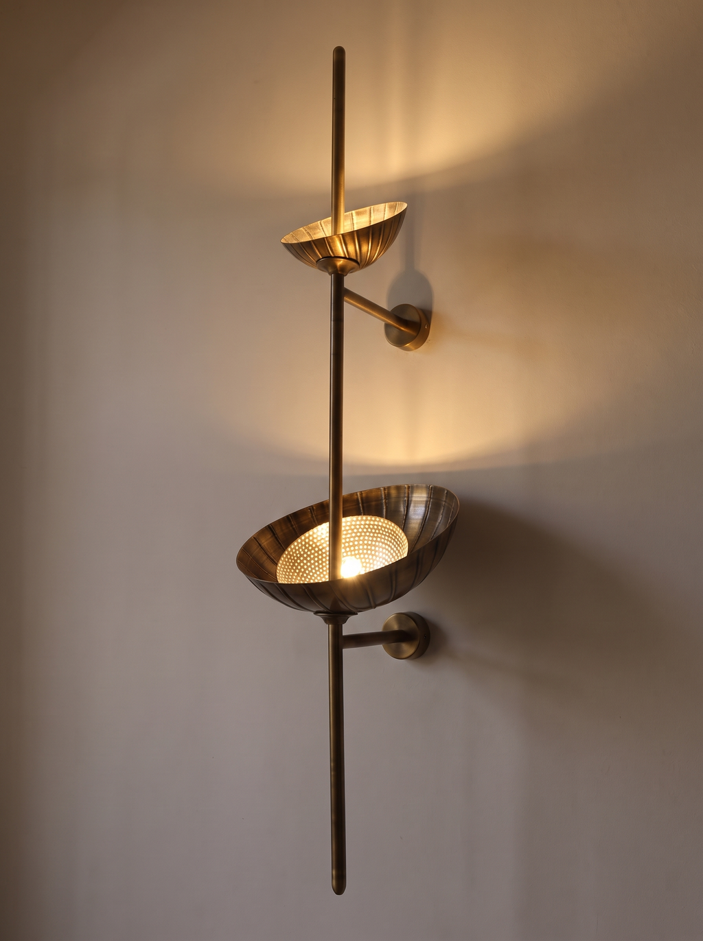 Stambha Etched Brass Wall Light switched on, both bowls glowing and washing the wall with warm uplight