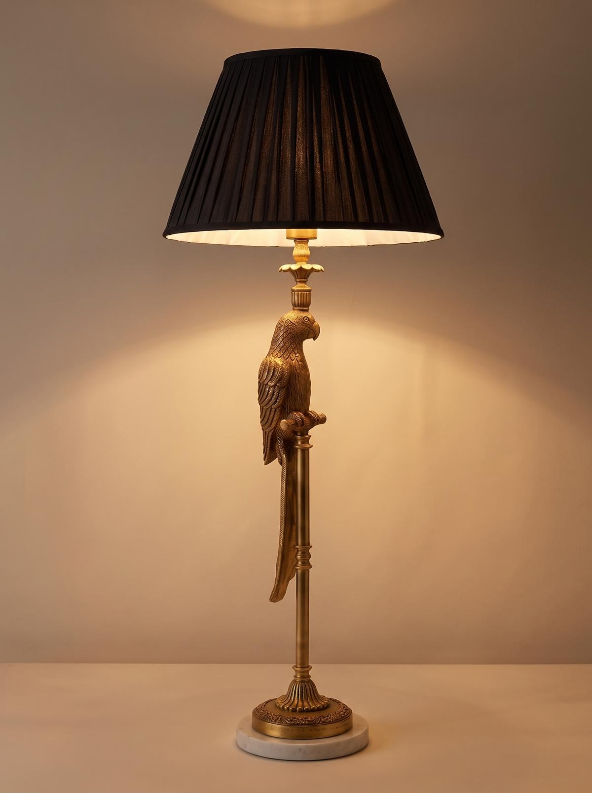 Brass Parrot Table Lamp switched on, warm light falling from the pleated shade onto the brass parrot